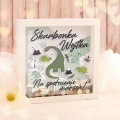 Drewniana skarbonka w kształcie kwadratu z szybką na przodzie. Na niej malownicza grafika dinozaurów, imienna dedykacja i napis "Na spełnienie marzeń!".