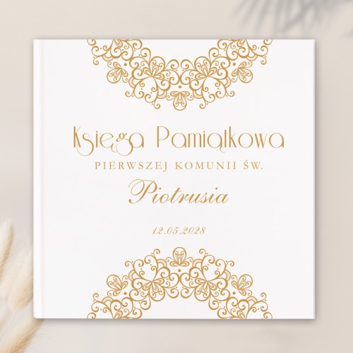 Biała księga pamiątkowa. W środku złoty napis Księga pamiątkowa Pierwszej Komunii Św. (imię dziecka); wraz z datą. U góry i dołu złoty ażurowy wzór w kształcie półkola.