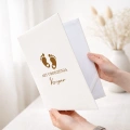 Personalizowana teczka na akt urodzenia dziecka ma elegancką białą okładkę ze złotymi stópkami i imieniem.