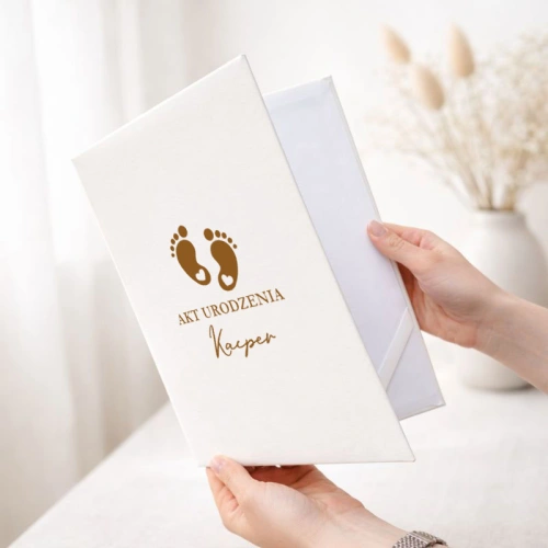 Personalizowana teczka na akt urodzenia dziecka ma elegancką białą okładkę ze złotymi stópkami i imieniem.