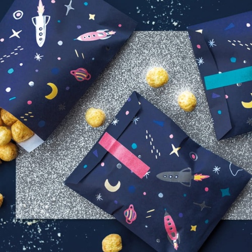 Torebki papierowe w granatowym kolorze ozdobione grafikami w kolorowe planety i kosmiczne rakiety. Dodatkowo uzupełnione ozdobnymi naklejkami.