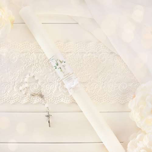 Biała świeca dekorowana grafiką z zielonymi listkami i białym krzyżem na ich tle. Imię dziecka umieszczone na świecy Dekorowana białym stroikiem.