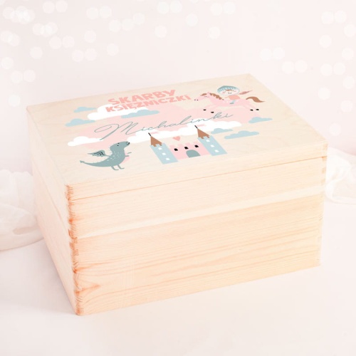 Duża skrzynia wykonana z jasnego naturalnego drewna. Wieko zdobione grafiką zamku, rycerza i smoka. Na pastelowym tle umieszczone jest imię dziecka.