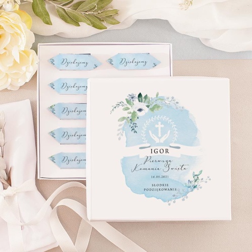 Komunijna bombonierka w kształcie kwadratu. W środku niebieskie krówki, na wierzchniej etykiecie akwarelowe  zdobienia z kwiatami i personalizowana dedykacja.