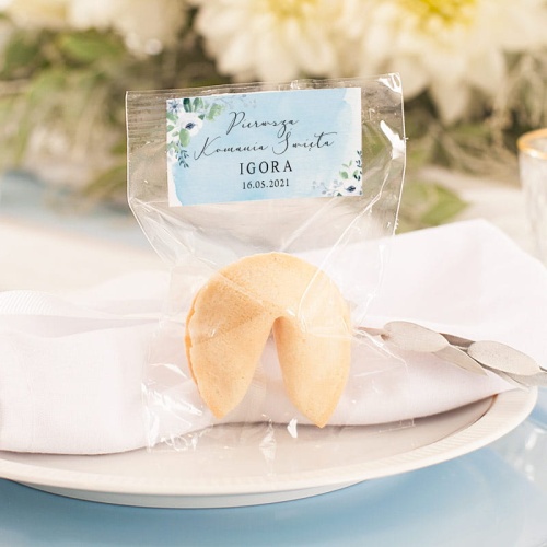 Ciasteczko z wróżbą w celofanowym woreczku z etykietą na górze. Na naklejce jest błękitna akwarela z kwiatkami, imienna dedykacja oraz data komunii.